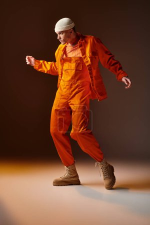 Foto de Hombre con estilo en mono naranja y chaqueta, gorro beige posando sobre fondo marrón - Imagen libre de derechos
