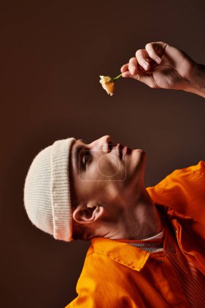 Vertical image of man in orange outfit wearing beige beanie holding flower in hand