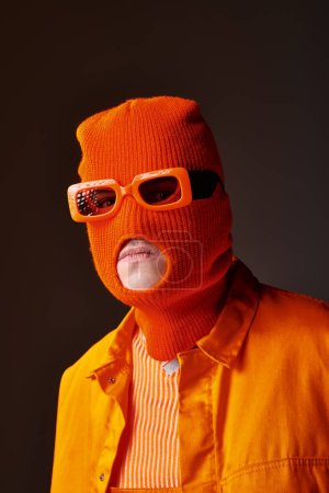 Stylischer Typ im orangefarbenen Outfit mit Sturmhaube und orangefarbener Sonnenbrille vor braunem Hintergrund