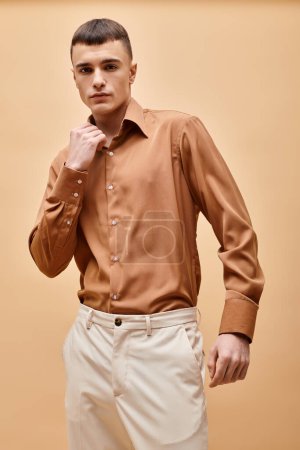 Portrait of stylish handsome man in beige shirt touching collar on peachy beige background