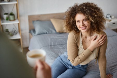 curly young woman smiling woman looking at boyfriend with coffee cup, morning couple time at bedroom