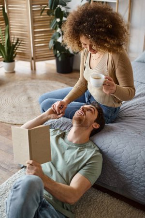 Cozy chat over coffee and book in bed, curly woman and brunette man enjoying each other's company