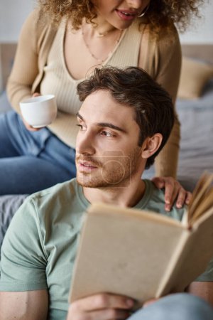 Homme brune détendu tenant le livre et regardant loin, étreint par sa petite amie, couple de temps matin