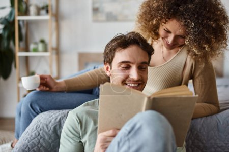 Smiling brunette man reading book and looking away, hugged by his girlfriend, morning couple time