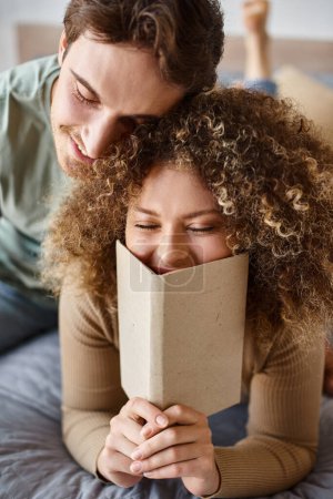 Téléchargez les photos : Doux matin embrasser de brune homme comme frisée jeune femme lit son livre dans le lit - en image libre de droit