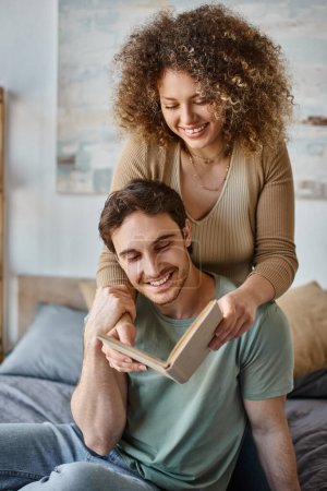 curly young woman lovingly hugs brunette man reading book in their cozy bedroom