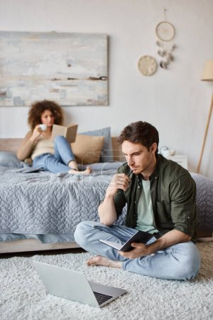 Téléchargez les photos : Un couple passe du temps à la maison. Femme lecture livre sur le lit et l'homme prend des notes assis sur le sol - en image libre de droit
