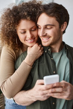 curly young woman kissing her brunette boyfriend, guy holding phone with smile and closed eyes