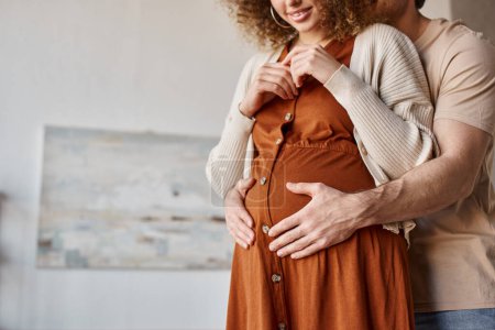 Cropped view of man hugging his pregnant wife from behind standing at home waiting for baby