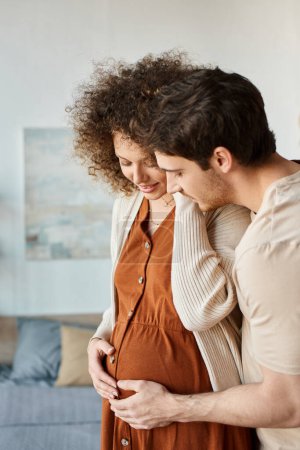 Precioso hombre feliz abrazando a su esposa embarazada por detrás de pie en casa esperando al bebé