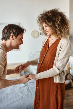 Cheerful man holding his pregnant wife hand and talking to their baby in belly of future mom