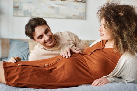 Téléchargez les photos : Femme enceinte avec mari dans la chambre, couchée au lit, homme touchant son ventre de femme - en image libre de droit
