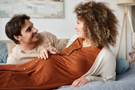 Pregnant woman with husband lying in bed, man touching his belly of wife and smiling