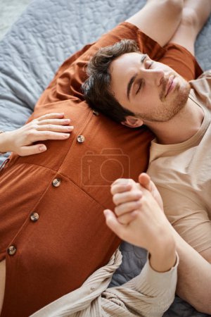 Pregnant woman with husband lying in bed, man resting near his belly of wife and smiling