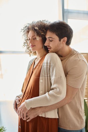 Expecting baby couple, man hugging his pregnant wife from behind standing near window