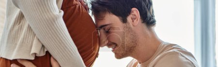 Foto de Vista lateral del sonriente hombre feliz esperando al bebé abrazando el vientre de la esposa con los ojos cerrados, pancarta - Imagen libre de derechos
