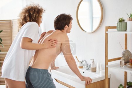 Photo for Back view of curly young woman and brunette man in bathroom looking in mirror, enjoying the morning - Royalty Free Image