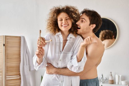 Téléchargez les photos : Jeune homme étreignant sa petite amie par derrière et riant tout en brossant les dents dans la salle de bain - en image libre de droit