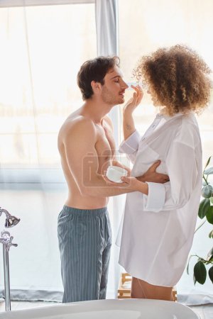 Foto de Mujer aplicando crema a su nariz de hombre en el baño y sonriendo. Vinculación de pareja por la mañana - Imagen libre de derechos