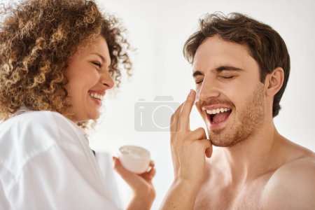 Téléchargez les photos : Femme appliquant de la crème à son homme dans la salle de bain et riant ensemble, heureux couple joyeux - en image libre de droit