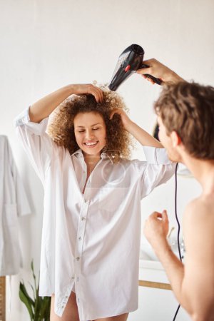 Foto de Feliz hombre secando su novia pelo rizado con secador de pelo por la mañana y riendo - Imagen libre de derechos