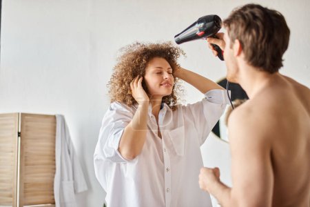 Téléchargez les photos : Homme séchant sa petite amie cheveux bouclés avec sèche-cheveux le matin, moment de liaison - en image libre de droit