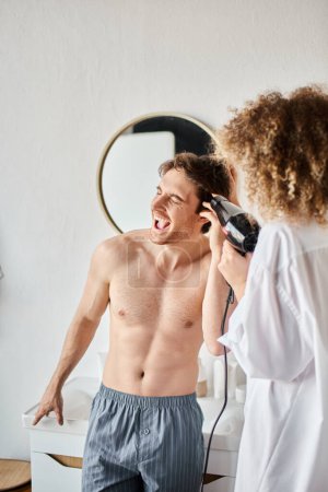Téléchargez les photos : Homme riant pendant que sa petite amie sèche ses cheveux avec sèche-cheveux le matin - en image libre de droit