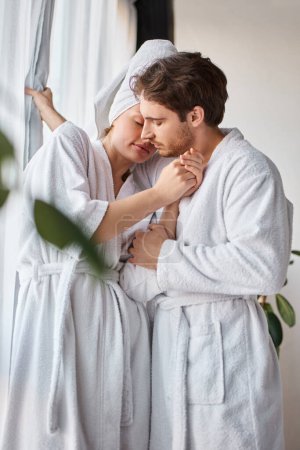 Couple in love wearing bathrobes hugging each other and having loving moment near window