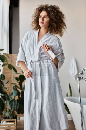 Beautiful curly young woman  wearing white robe in bathroom tying the belt and looking away