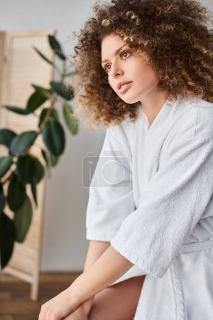 Lockige schöne junge Frau in weißem Gewand sitzt im Badezimmer und schaut weg
