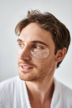 Porträt eines hübschen brünetten Mannes mit Augenklappen, der im Badezimmer ein weißes Gewand trägt und wegschaut