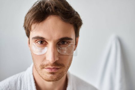 Porträt eines brünetten Mannes mit Augenklappen, der im Badezimmer einen weißen Mantel trägt und in die Kamera blickt