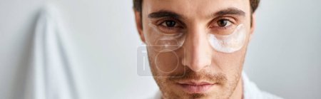 Téléchargez les photos : Portrait d'homme brune avec des patchs pour les yeux portant une robe blanche dans la salle de bain en regardant la caméra, bannière - en image libre de droit