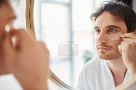 Porträt eines brünetten Mannes mit Augenklappen, der im Badezimmer einen weißen Mantel trägt und in den Spiegel schaut