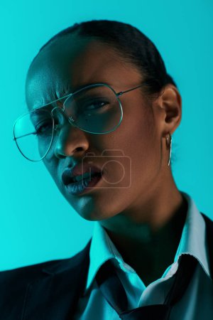 A young African American woman in glasses and a suit standing confidently in a studio setting