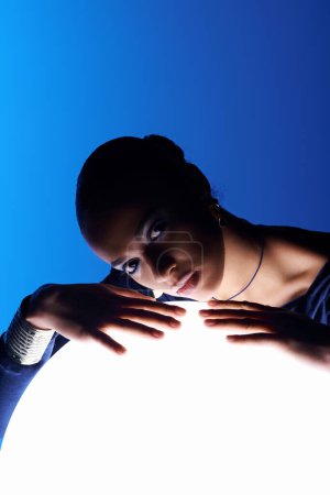 A young African American woman holds a glowing light ball in front of her face in a studio setting.
