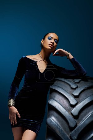 A young African American woman stands confidently next to a colossal tire in a studio setting.