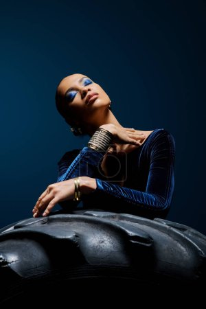 Joven mujer afroamericana con un vestido azul se sienta con gracia en la parte superior de un neumático en un entorno de estudio.