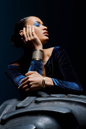 A young African American woman wearing blue makeup leans against a tire in a studio setting.