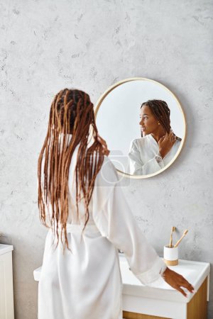 A woman with dreadlocks stands in front of a mirror in a modern bathroom, examining her beauty in a bathrobe.