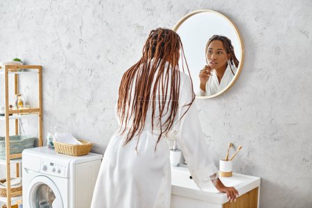 An African American woman in a bath robe and afro braids brushes her teeth in front of a mirror in a modern bathroom.