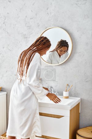 Une femme afro-américaine avec des tresses afro se tient dans sa salle de bain moderne, s'engageant dans des rituels de beauté et d'hygiène.