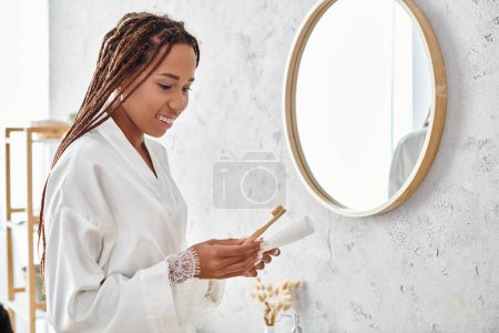 Eine afroamerikanische Frau mit Afro-Zöpfen steht in einem modernen Badezimmer und hält im Bademantel eine Bürste in der Hand.