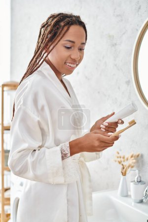 An African American woman in a bathrobe stands in front of a modern bathroom sink, engaging in a beauty and hygiene routine.