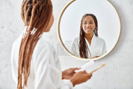 Eine afroamerikanische Frau mit Afro-Zöpfen steht in einem modernen Badezimmer vor einem Spiegel und trägt einen Bademantel.