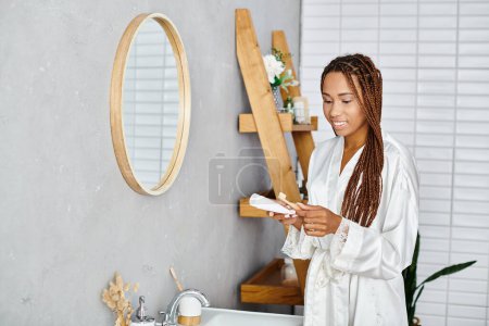 Foto de Una mujer afroamericana con trenzas afro se encuentra en su moderno baño, centrándose en las rutinas de belleza e higiene en el fregadero. - Imagen libre de derechos