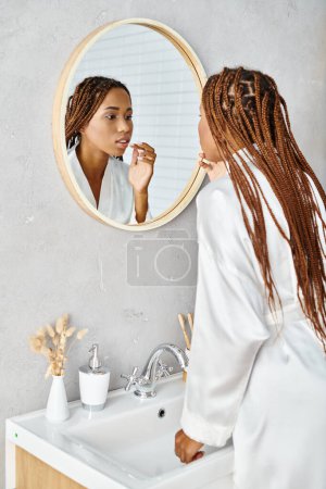 An African American woman with afro braids brushes her teeth in a modern bathroom mirror while wearing a bath robe.