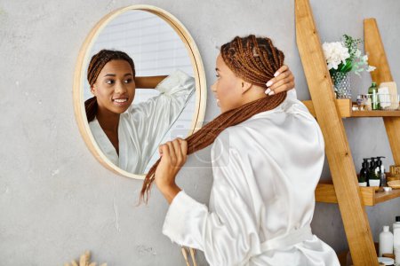 A stylish African American woman with afro braids brushes her hair in front of a mirror in a modern bathroom.