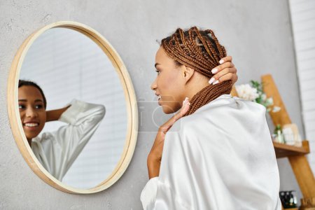Une femme avec des tresses afro se tient devant un miroir dans une salle de bain moderne, se brossant les cheveux dans un peignoir de bain.
