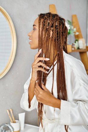 Eine afroamerikanische Frau mit Afro-Zöpfen bürstet sich in einem modernen Badezimmer die Haare, während sie einen Bademantel trägt.